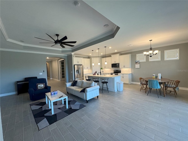 living room with a raised ceiling, crown molding, sink, and ceiling fan with notable chandelier