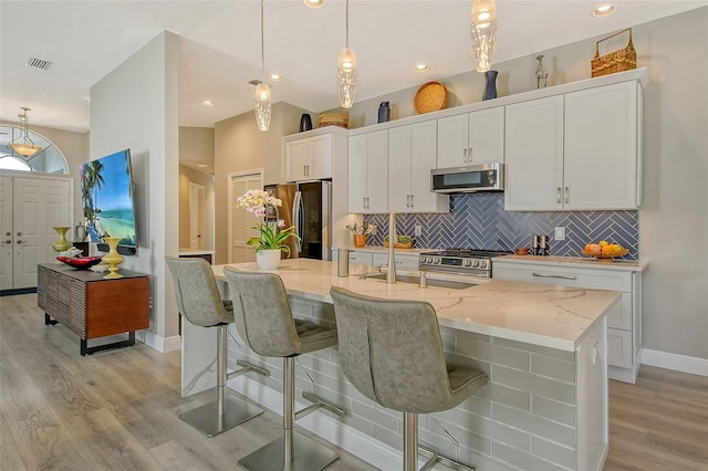 kitchen with white cabinetry, stainless steel appliances, light stone countertops, a center island with sink, and decorative light fixtures