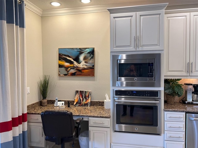 kitchen featuring stainless steel appliances, ornamental molding, white cabinets, built in study area, and dark stone countertops