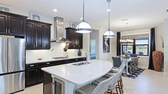 kitchen with appliances with stainless steel finishes, decorative light fixtures, beverage cooler, a kitchen island with sink, and wall chimney range hood