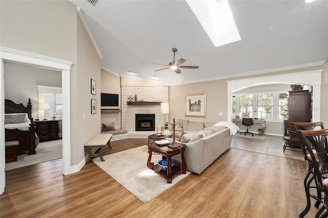 living area featuring a ceiling fan, lofted ceiling with skylight, light wood-style flooring, ornamental molding, and a brick fireplace