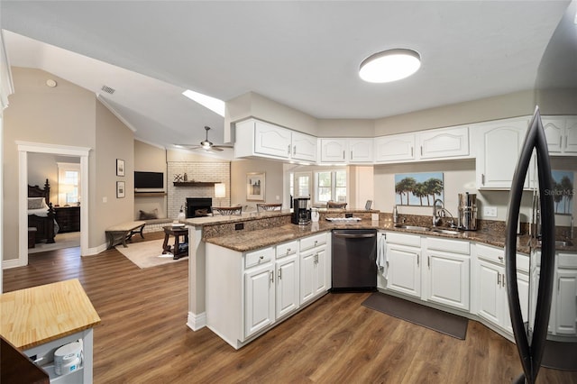 kitchen featuring dark stone counters, a peninsula, stainless steel dishwasher, a brick fireplace, and a sink