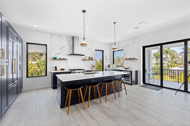 kitchen featuring a breakfast bar, wine cooler, a center island with sink, decorative light fixtures, and wall chimney exhaust hood