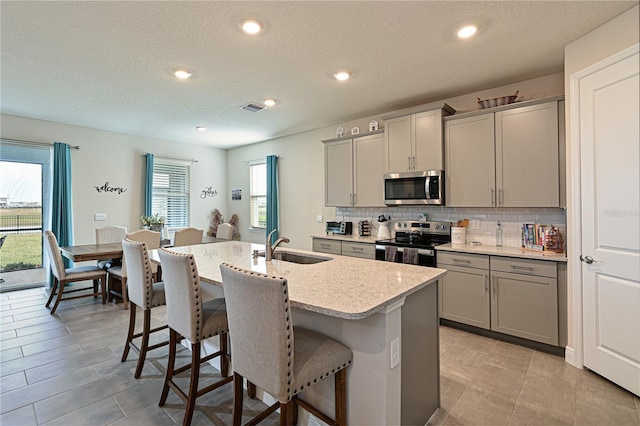 kitchen featuring gray cabinets, sink, backsplash, stainless steel appliances, and a center island with sink