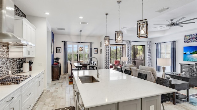 kitchen with a breakfast bar area, a kitchen island with sink, hanging light fixtures, white cabinets, and wall chimney exhaust hood