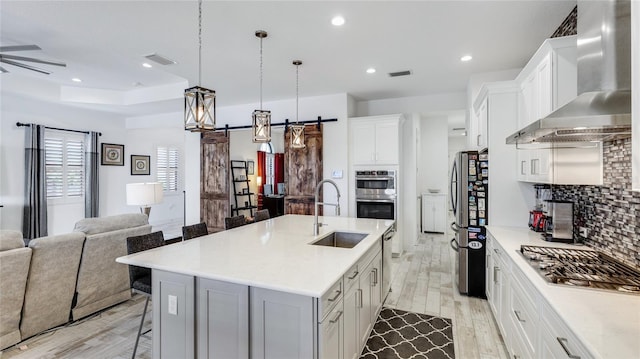 kitchen featuring sink, a kitchen island with sink, stainless steel appliances, a barn door, and wall chimney exhaust hood