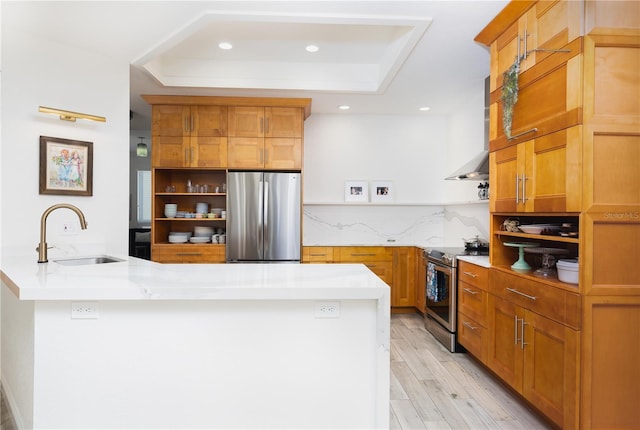 kitchen with sink, stainless steel appliances, a tray ceiling, wall chimney exhaust hood, and light wood-type flooring