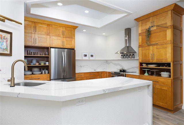 kitchen featuring sink, light stone counters, a tray ceiling, stainless steel appliances, and wall chimney exhaust hood