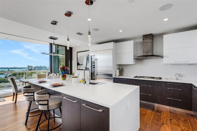 kitchen with built in fridge, decorative light fixtures, an island with sink, white cabinetry, and wall chimney exhaust hood