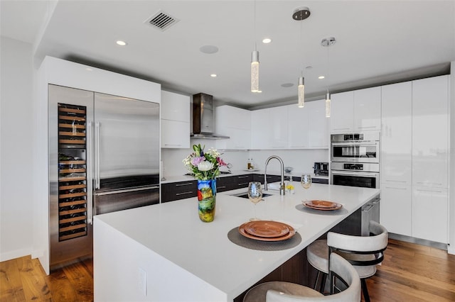 kitchen with appliances with stainless steel finishes, pendant lighting, a center island with sink, and wall chimney exhaust hood