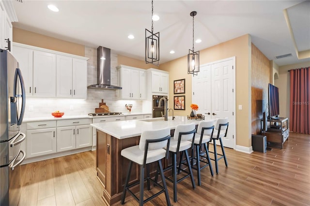 kitchen with wall chimney exhaust hood, white cabinetry, decorative light fixtures, an island with sink, and stainless steel appliances