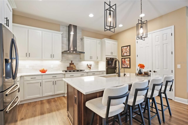 kitchen featuring a kitchen island with sink, white cabinetry, stainless steel appliances, decorative light fixtures, and wall chimney exhaust hood