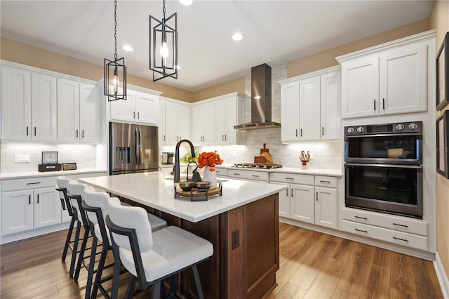 kitchen featuring wall chimney range hood, white cabinetry, hanging light fixtures, stainless steel appliances, and an island with sink