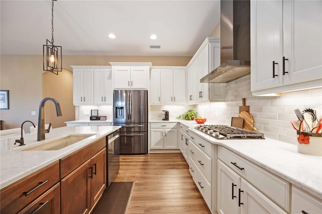 kitchen featuring wall chimney range hood, sink, stainless steel appliances, white cabinets, and decorative light fixtures