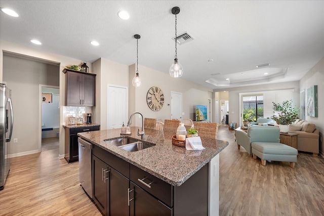 kitchen with sink, a kitchen island with sink, dark brown cabinets, a tray ceiling, and light stone countertops