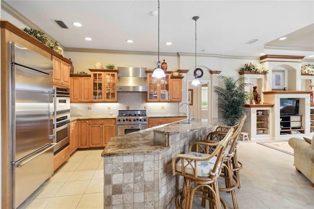 kitchen with wall chimney range hood, a breakfast bar, premium appliances, light stone countertops, and decorative light fixtures