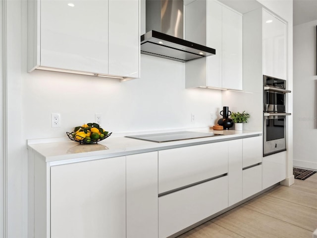 kitchen with double oven, white cabinets, wall chimney range hood, black electric cooktop, and light hardwood / wood-style flooring