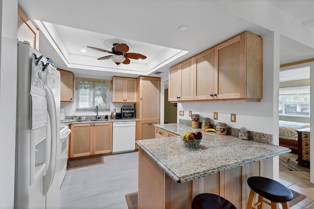 kitchen featuring a kitchen bar, light brown cabinetry, a tray ceiling, kitchen peninsula, and white appliances