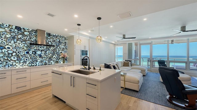 kitchen with a water view, decorative light fixtures, white cabinets, a kitchen island with sink, and wall chimney range hood