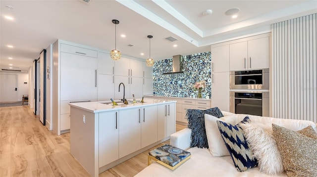 kitchen with pendant lighting, sink, wall chimney range hood, a kitchen island with sink, and white cabinetry