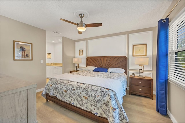 bedroom with ceiling fan, ensuite bath, light hardwood / wood-style flooring, and a textured ceiling