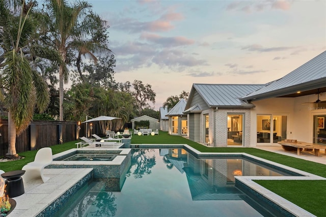 pool at dusk featuring a yard, a patio area, ceiling fan, and an in ground hot tub