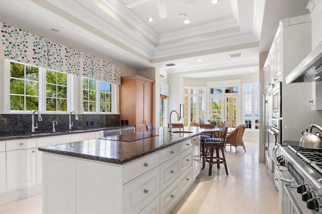 kitchen with dark stone countertops, wall chimney exhaust hood, an island with sink, and white cabinets