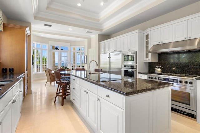 kitchen with white cabinetry, dark stone countertops, built in appliances, an island with sink, and a raised ceiling