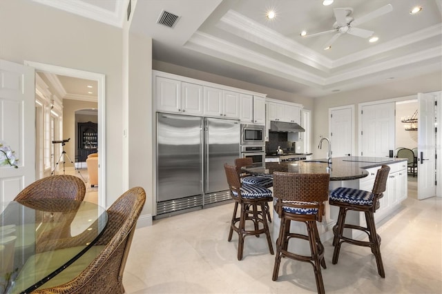 kitchen featuring sink, white cabinetry, built in appliances, a tray ceiling, and a center island with sink