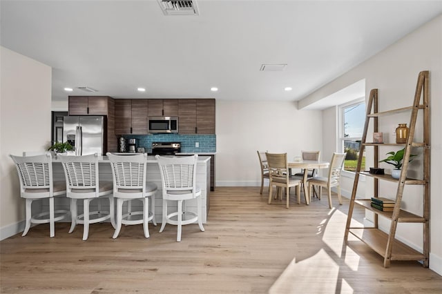 kitchen with a breakfast bar area, decorative backsplash, light hardwood / wood-style floors, dark brown cabinetry, and stainless steel appliances