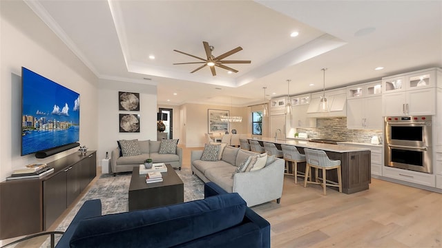 living room with ceiling fan, ornamental molding, a raised ceiling, and light wood-type flooring