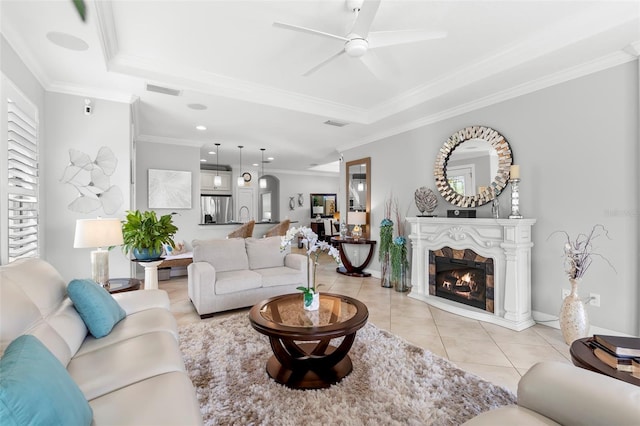 living area featuring a tray ceiling, light tile patterned floors, ornamental molding, a glass covered fireplace, and ceiling fan