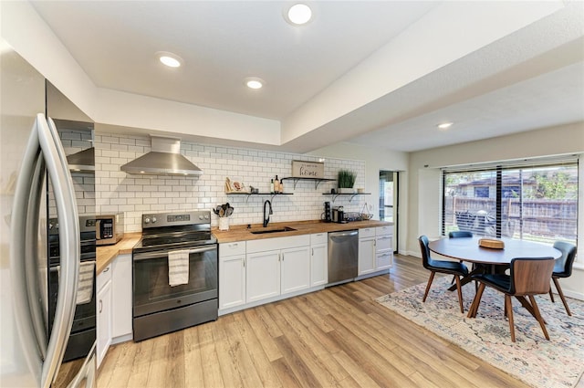 kitchen with a sink, wood counters, light wood-style floors, wall chimney range hood, and appliances with stainless steel finishes