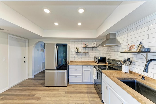 kitchen with light wood-style flooring, stainless steel appliances, butcher block counters, a sink, and wall chimney range hood