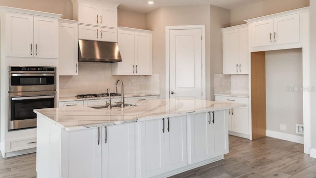 kitchen with light stone counters, stainless steel appliances, an island with sink, and white cabinets