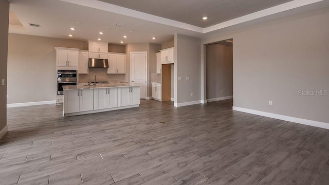 kitchen featuring light hardwood / wood-style flooring, white cabinetry, a center island with sink, decorative backsplash, and stainless steel double oven