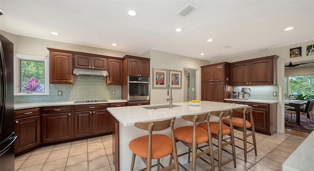 kitchen featuring sink, light tile patterned floors, a center island with sink, a kitchen bar, and black electric cooktop