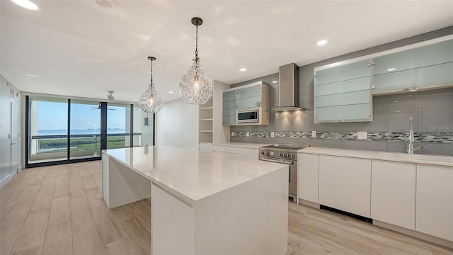 kitchen with light countertops, appliances with stainless steel finishes, a center island, wall chimney exhaust hood, and glass insert cabinets