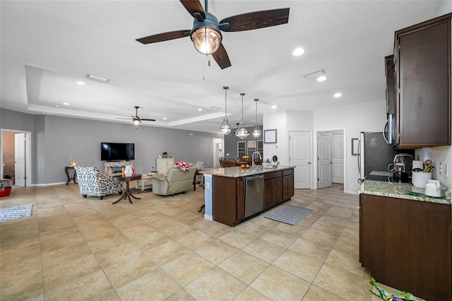 kitchen with decorative light fixtures, dishwasher, a kitchen island with sink, light stone counters, and a raised ceiling