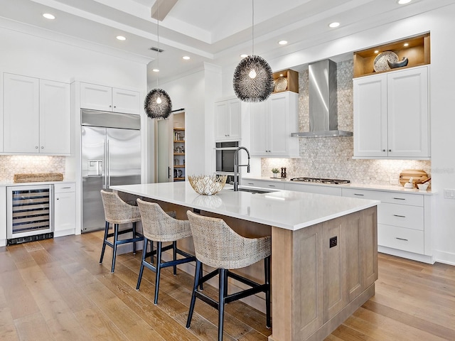 kitchen with wine cooler, built in fridge, light countertops, wall chimney exhaust hood, and a center island with sink