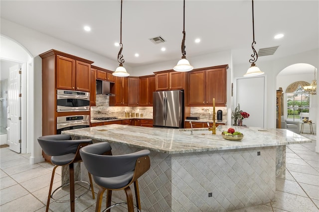 kitchen with arched walkways, wall chimney exhaust hood, appliances with stainless steel finishes, a kitchen bar, and pendant lighting