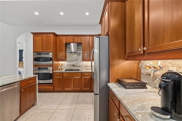 kitchen featuring brown cabinetry, wall chimney exhaust hood, light stone counters, stainless steel appliances, and backsplash