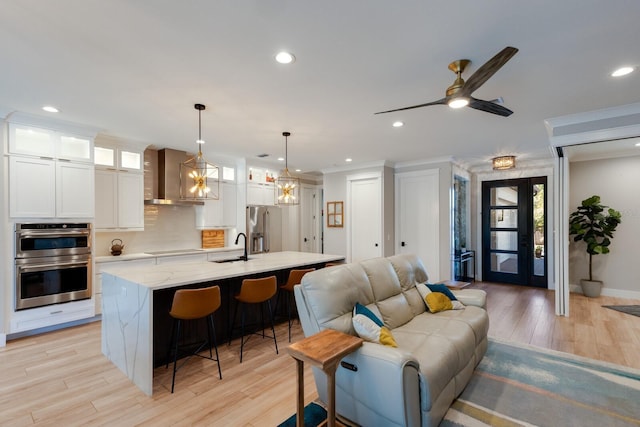 kitchen featuring wall chimney exhaust hood, hanging light fixtures, stainless steel appliances, a kitchen island with sink, and white cabinets