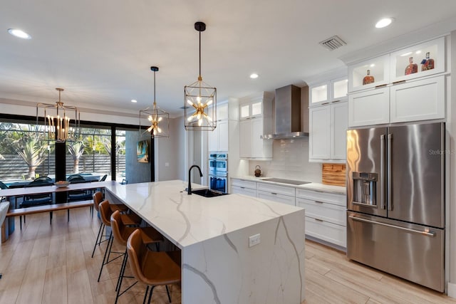 kitchen with wall chimney range hood, sink, stainless steel appliances, an island with sink, and white cabinets