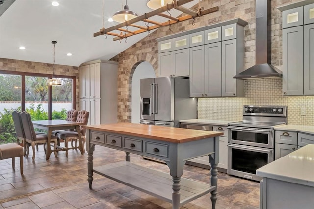 kitchen featuring wood counters, appliances with stainless steel finishes, vaulted ceiling, backsplash, and wall chimney exhaust hood