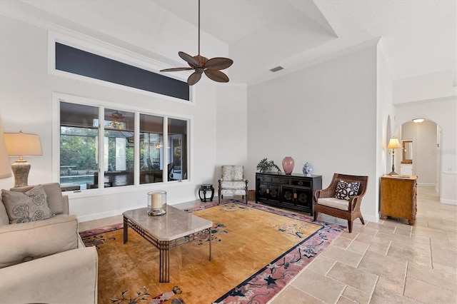 living area with arched walkways, stone tile floors, a raised ceiling, ceiling fan, and baseboards