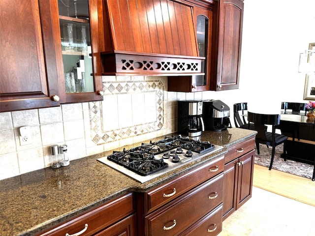 kitchen with stainless steel gas cooktop, backsplash, glass insert cabinets, wall chimney range hood, and dark stone countertops