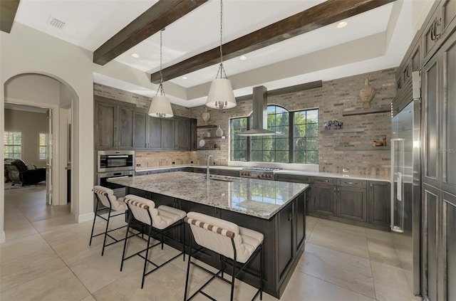 kitchen with arched walkways, a center island with sink, open shelves, wall chimney range hood, and a kitchen breakfast bar