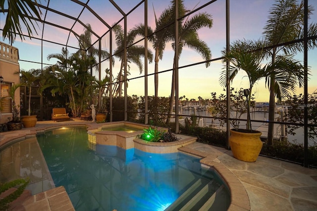 pool at dusk with an outdoor pool, a patio area, glass enclosure, and a jacuzzi