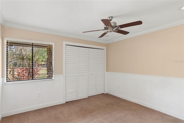 unfurnished bedroom featuring wainscoting, ornamental molding, carpet floors, a closet, and ceiling fan